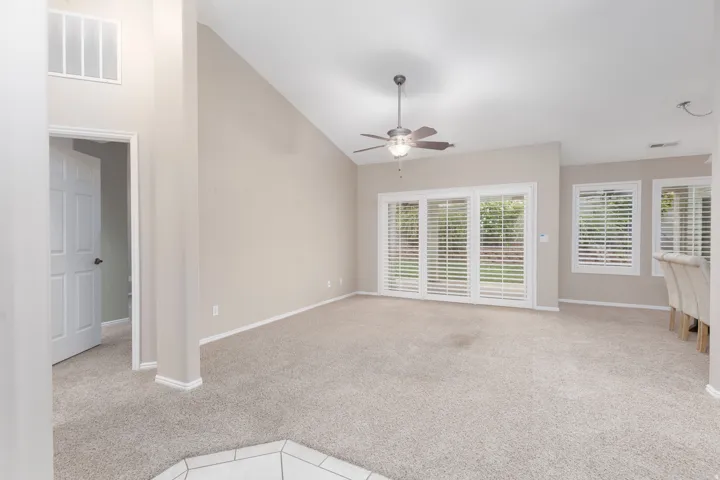 Vaulted ceiling in a carpeted room featuring a ceiling fan, sliding glass doors with plantation shutters, and additional windows with plantation shutters