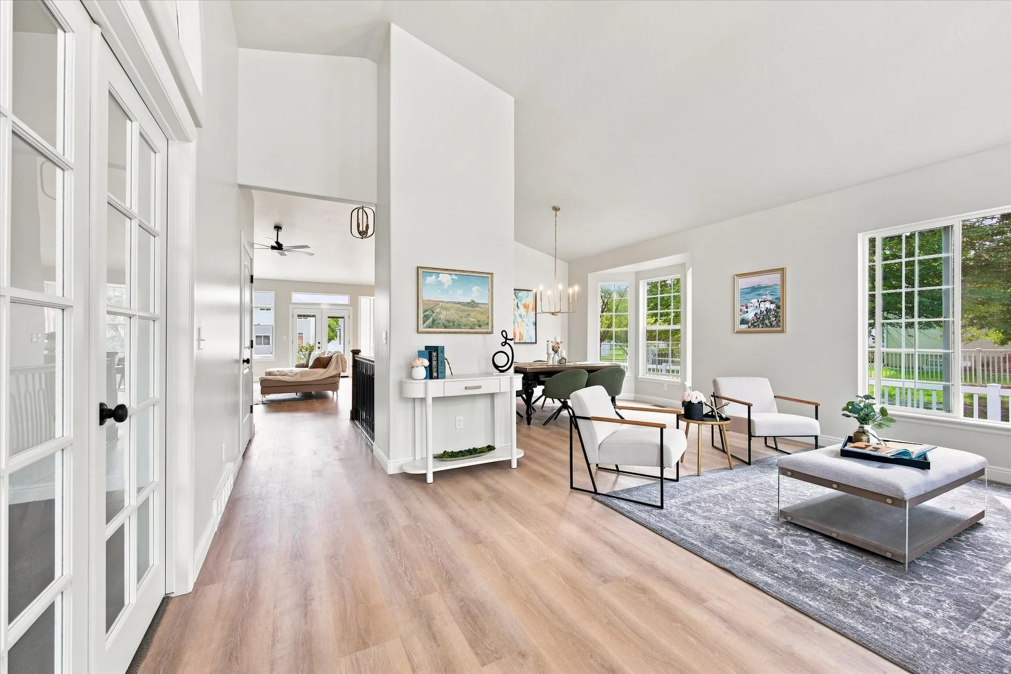 Bright, open-concept living area featuring wide-plank wood-finish flooring, abundant natural light from multiple multi-pane windows, a central kitchen island with white cabinetry, and a spacious great room with neutral wall finishes and area rugging