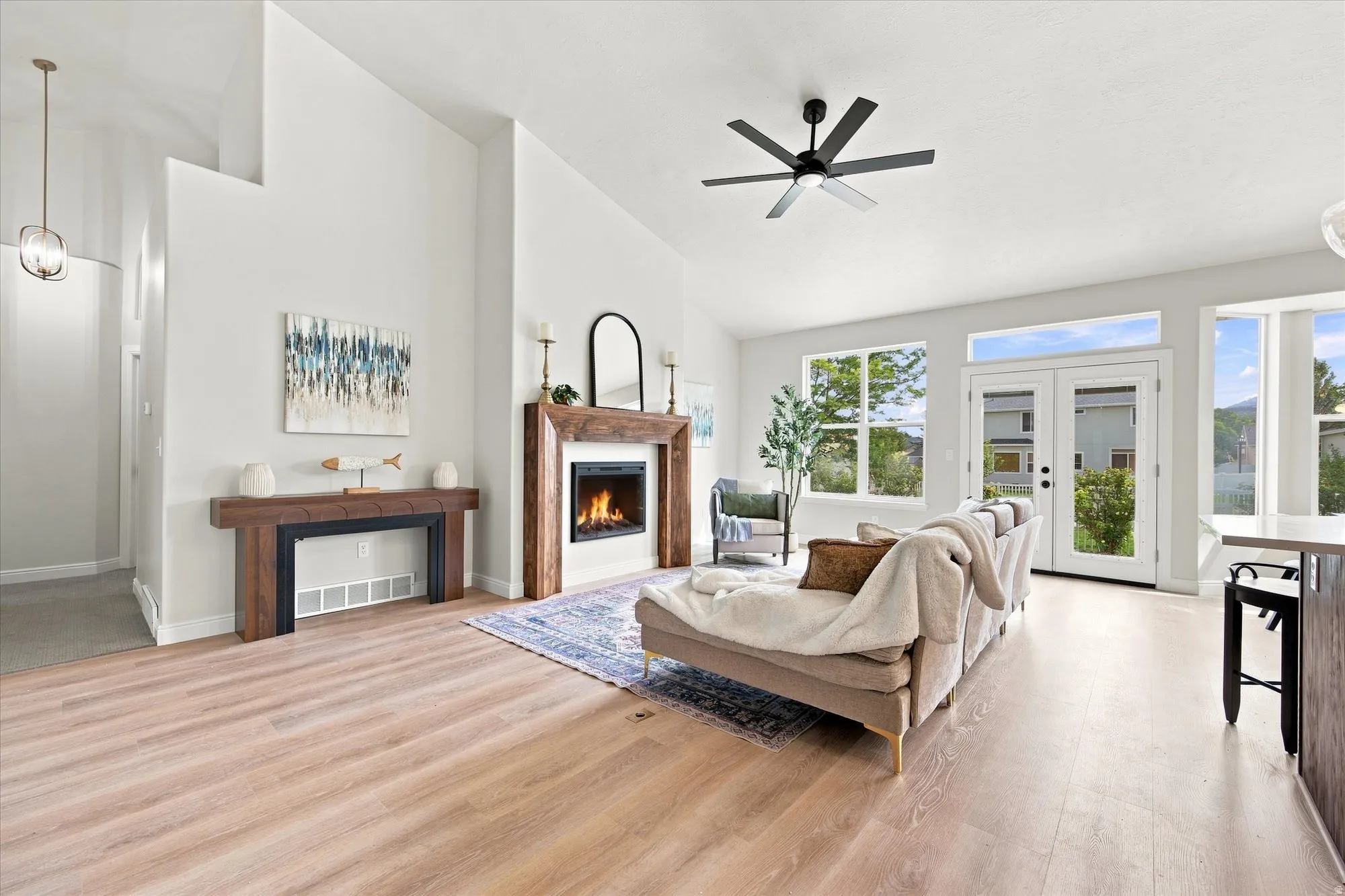 Spacious living area featuring wood-finish flooring, a contemporary fireplace with a wood surround, a modern ceiling fan, and abundant natural light through multiple windows and French doors