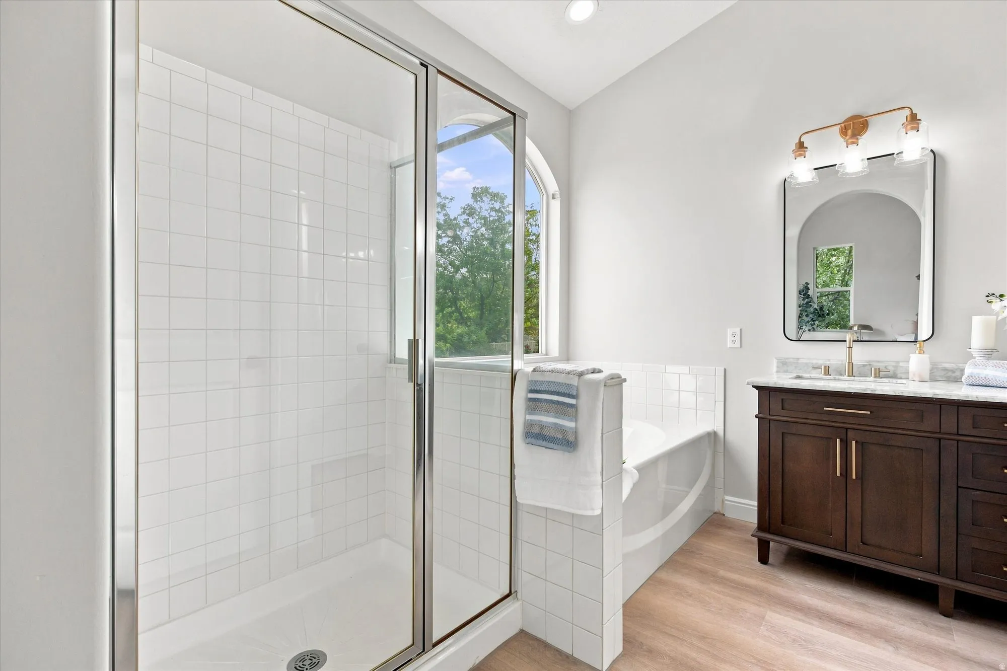 Glass-enclosed shower with white tiled walls, separate soaking tub, dark wood-finish vanity with countertop, rectangular mirror, and wood-finish flooring