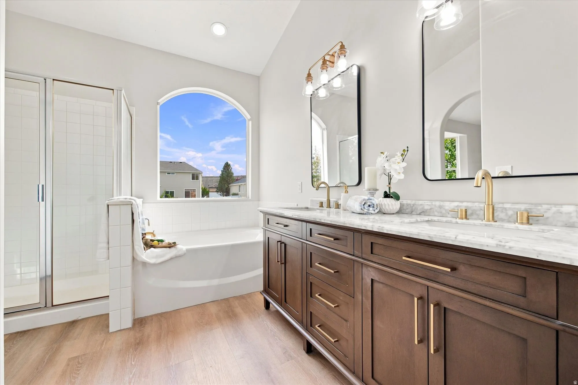 Dual vanity with stone countertops and brass fixtures, arched window, built-in soaking tub, and glass-enclosed shower