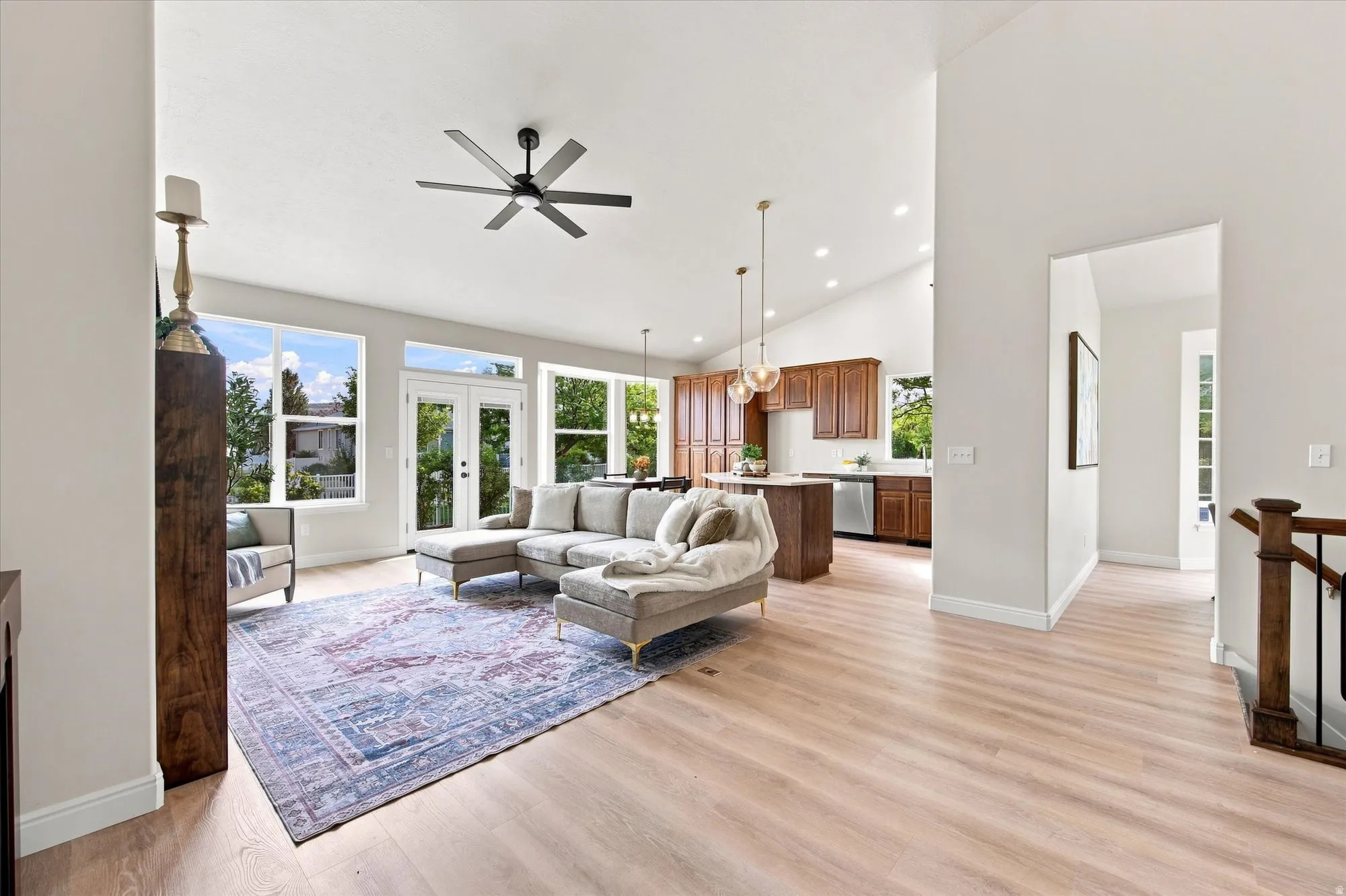 Bright open-concept living area showcasing soaring ceilings with ceiling fan, expansive windows with natural light, wood-finish flooring throughout, modern kitchen with cabinetry and pendant lighting, and a decorative area rug anchoring the seating zone