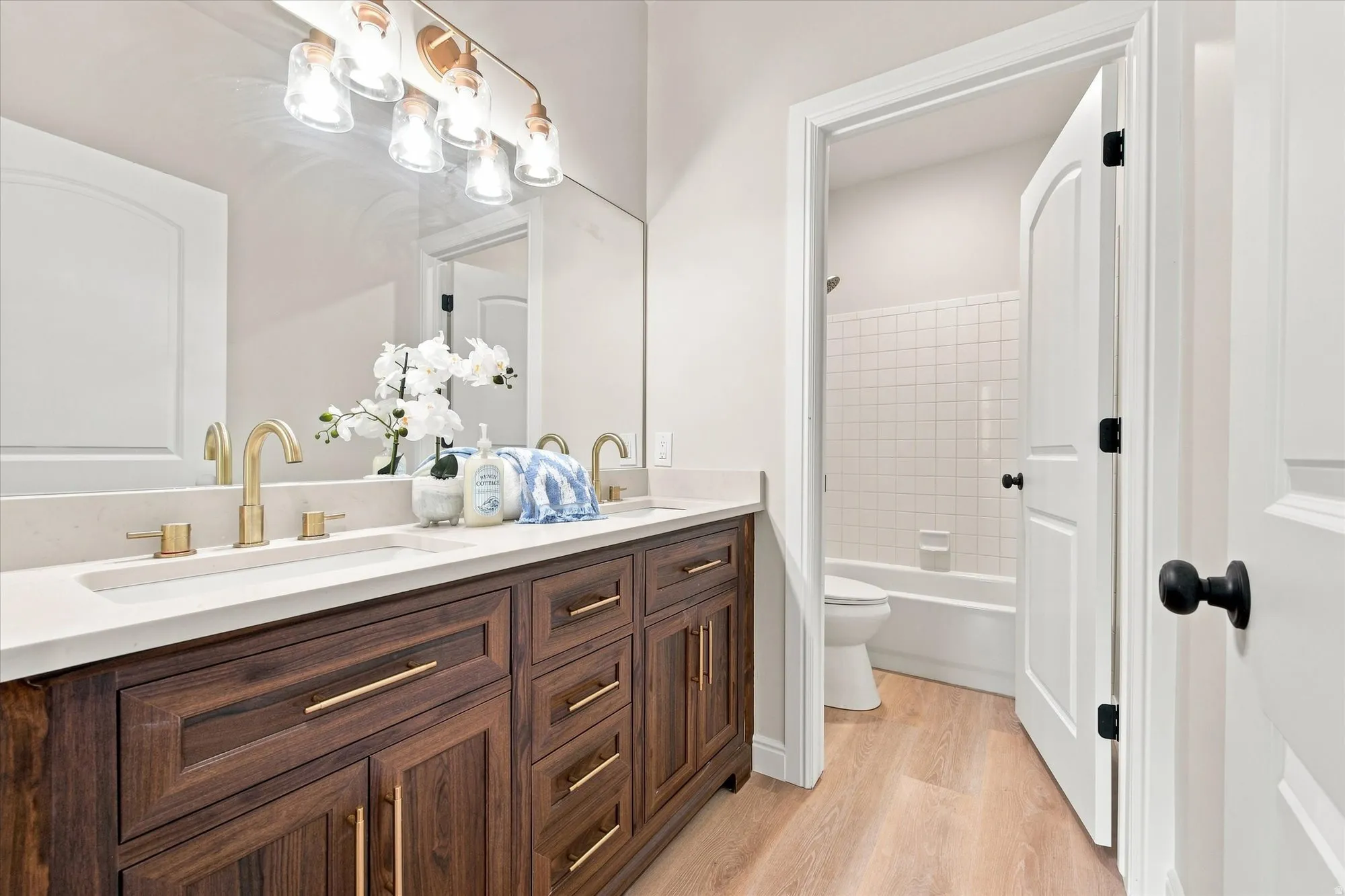 Dual vanity with integrated sinks, wood-finish cabinetry, gold-tone hardware, and an overhead vanity light