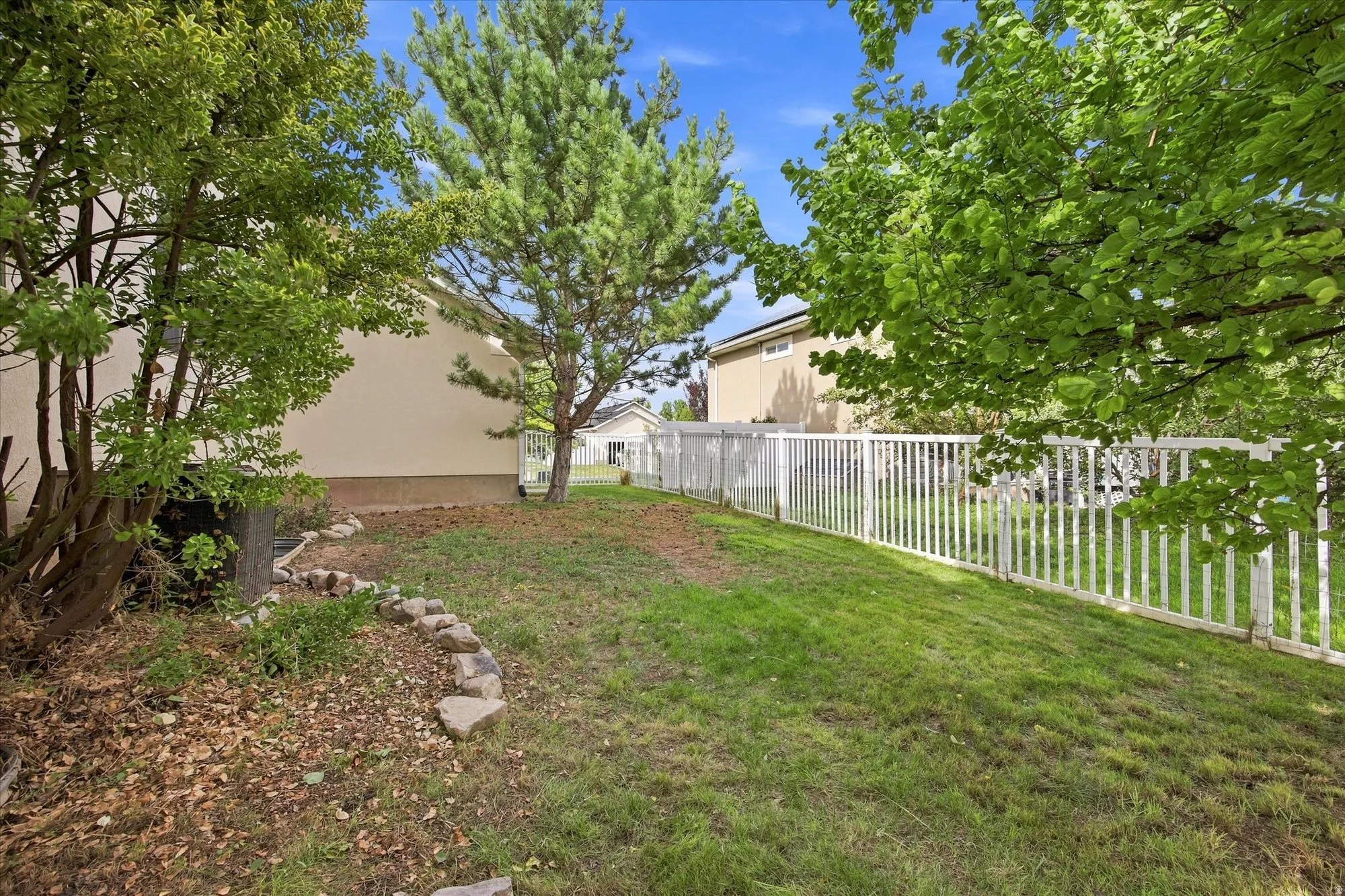 Landscaped yard featuring mature trees, a white picket fence, and a stone-lined garden bed