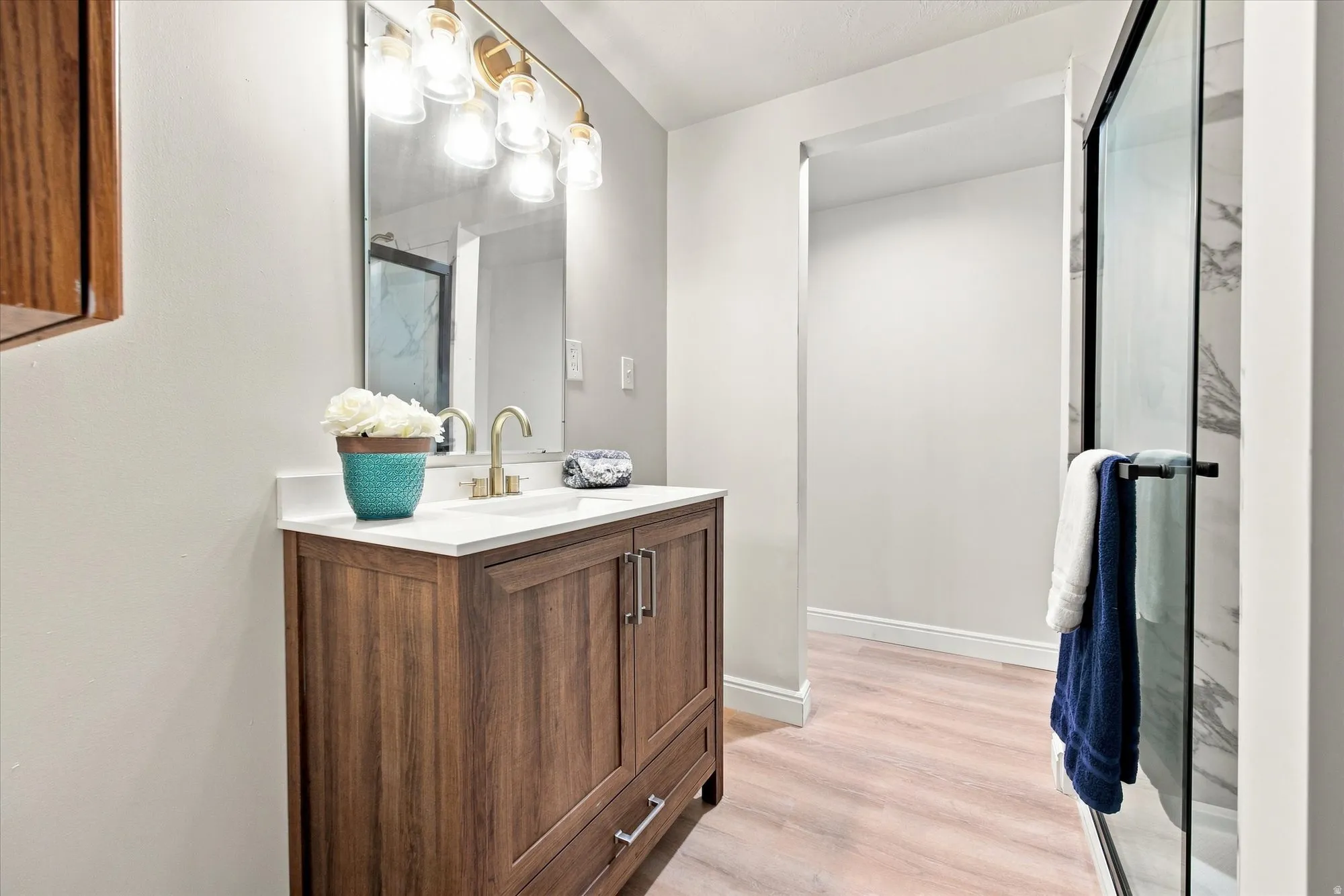 Wood-finish vanity with a white countertop, gold-tone faucet, framed mirror, and a glass-enclosed shower with a marble-patterned surround