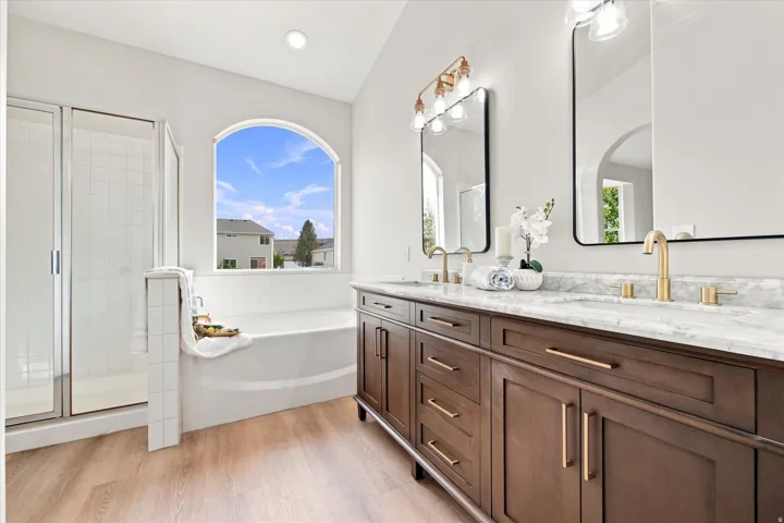 Dual vanity with stone countertops and brass fixtures, arched window, built-in soaking tub, and glass-enclosed shower