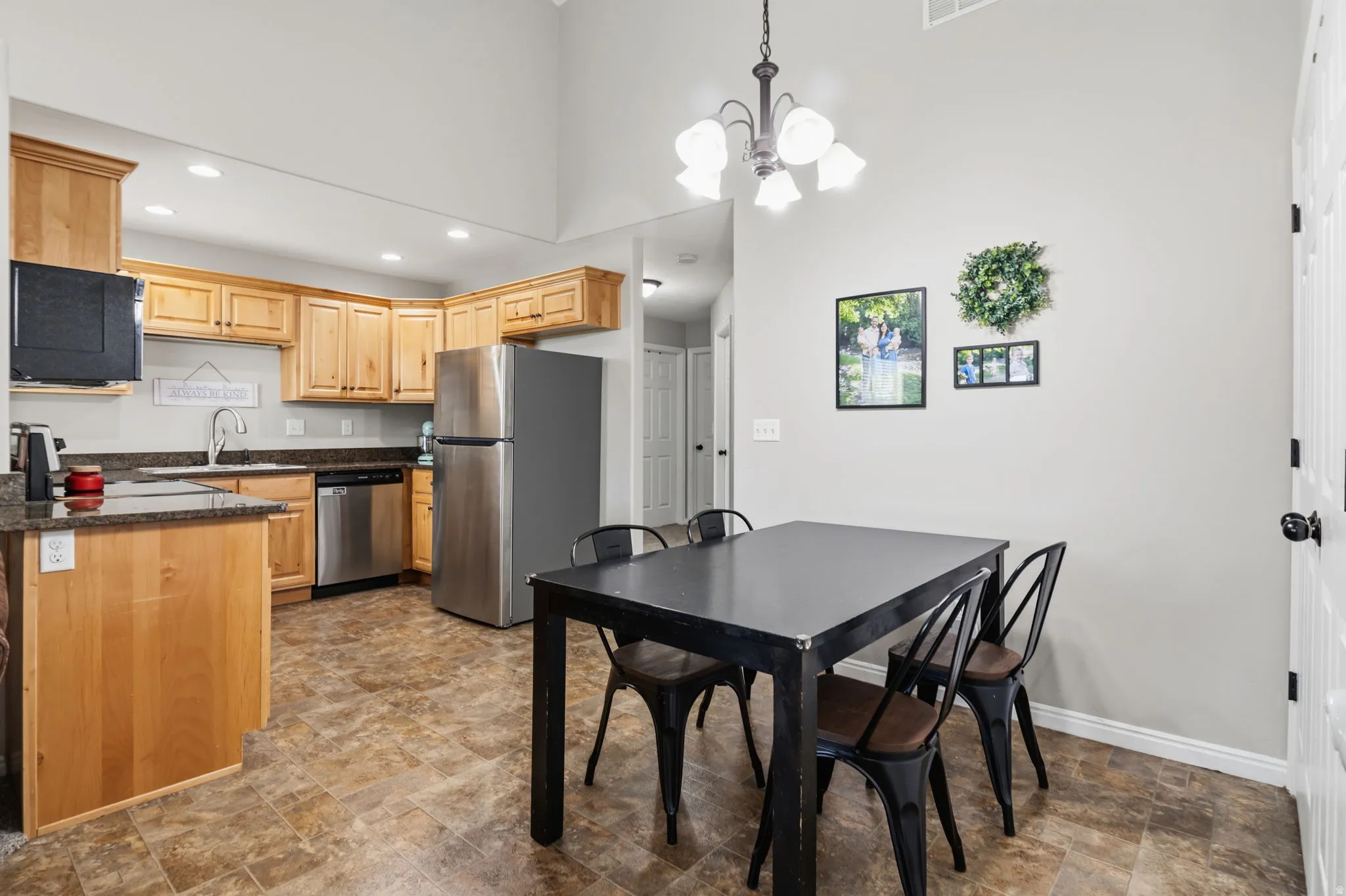 Open-concept kitchen and dining area featuring warm wood cabinetry, stainless steel appliances, stone-finish flooring, a black dining table with seating for four, and a pendant light fixture overhead