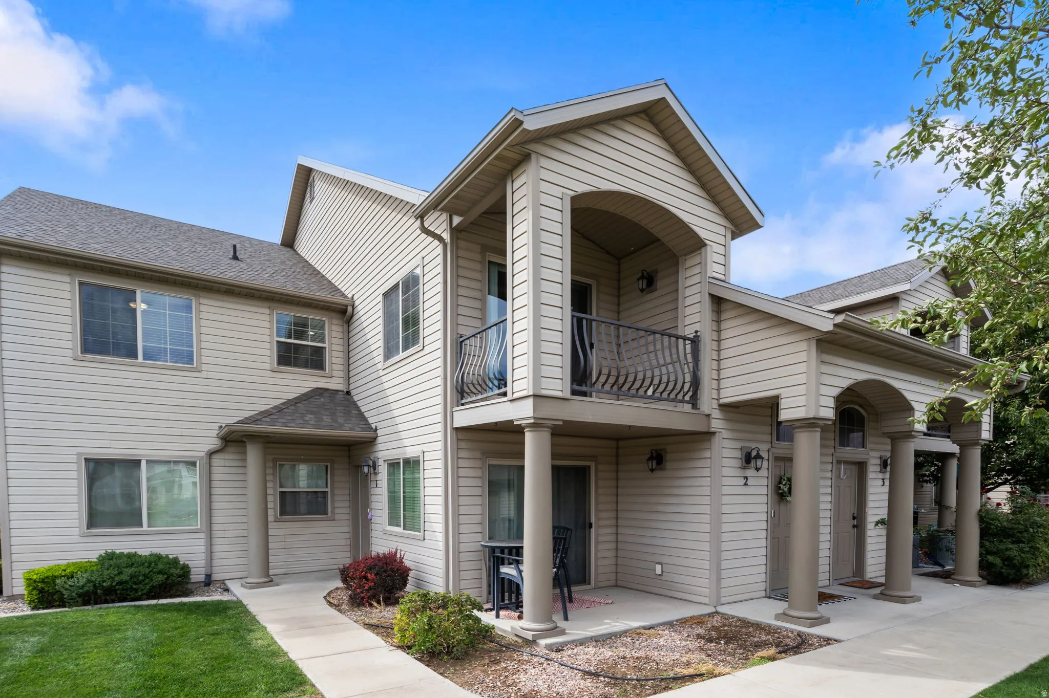Multi-unit exterior with light-colored siding, a second-story balcony with decorative railing, arched entryways, and columned porch supports