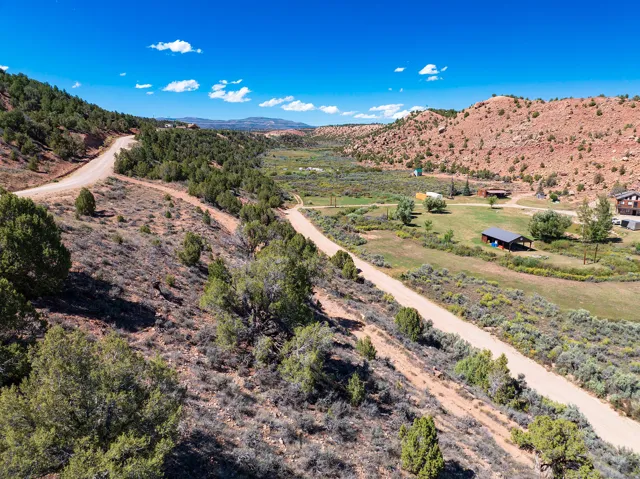 Expansive rural landscape featuring a winding dirt road, scattered evergreen trees, and distant red rock formations