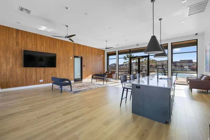 Spacious communal area featuring extensive wood-finish flooring, a full-height wood-paneled accent wall, a central kitchen island with a light-toned countertop, and large floor-to-ceiling windows