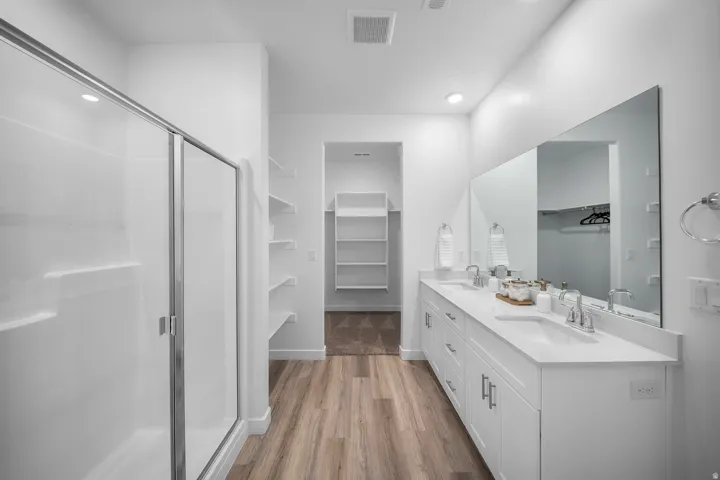 Wood-finish flooring with a white double vanity, large frameless mirror, and a walk-in shower with a sliding glass door