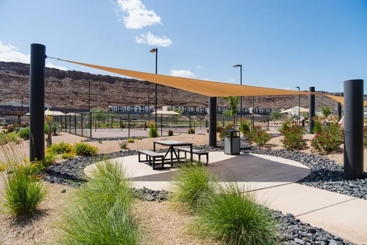 Outdoor recreational area featuring a shade sail, concrete patio, picnic table with benches, and fenced courts