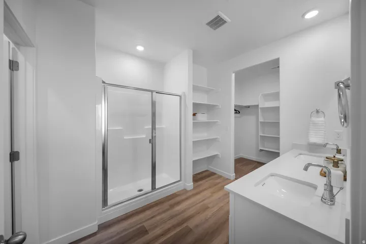 Walk-in shower with sliding glass doors, dual vanity with undermount sinks, wood-finish flooring, recessed lighting, and built-in shelving