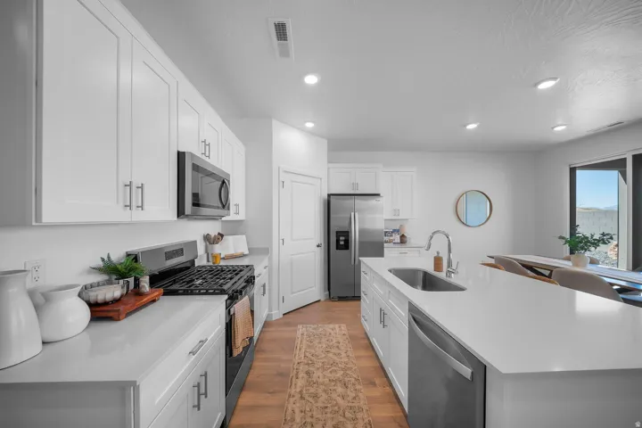 Modern kitchen featuring white shaker cabinetry, stainless steel appliances including a gas cooktop and under-counter microwave, quartz countertops, a large center island with integrated sink, recessed ceiling lighting, wood-finish flooring, and abundant