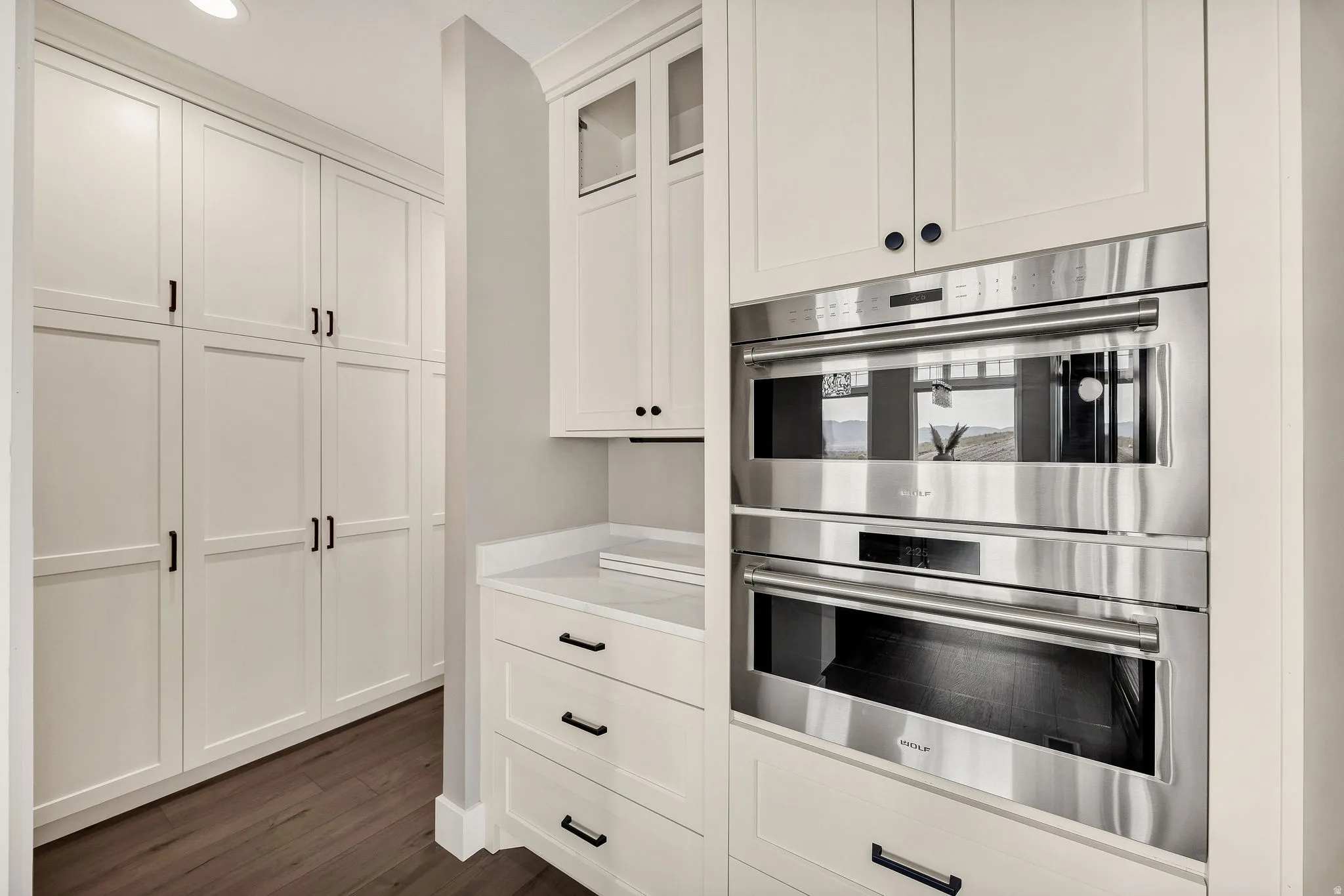 Built-in stainless steel double ovens, extensive white cabinetry with black hardware, white countertop, and wood-finish flooring