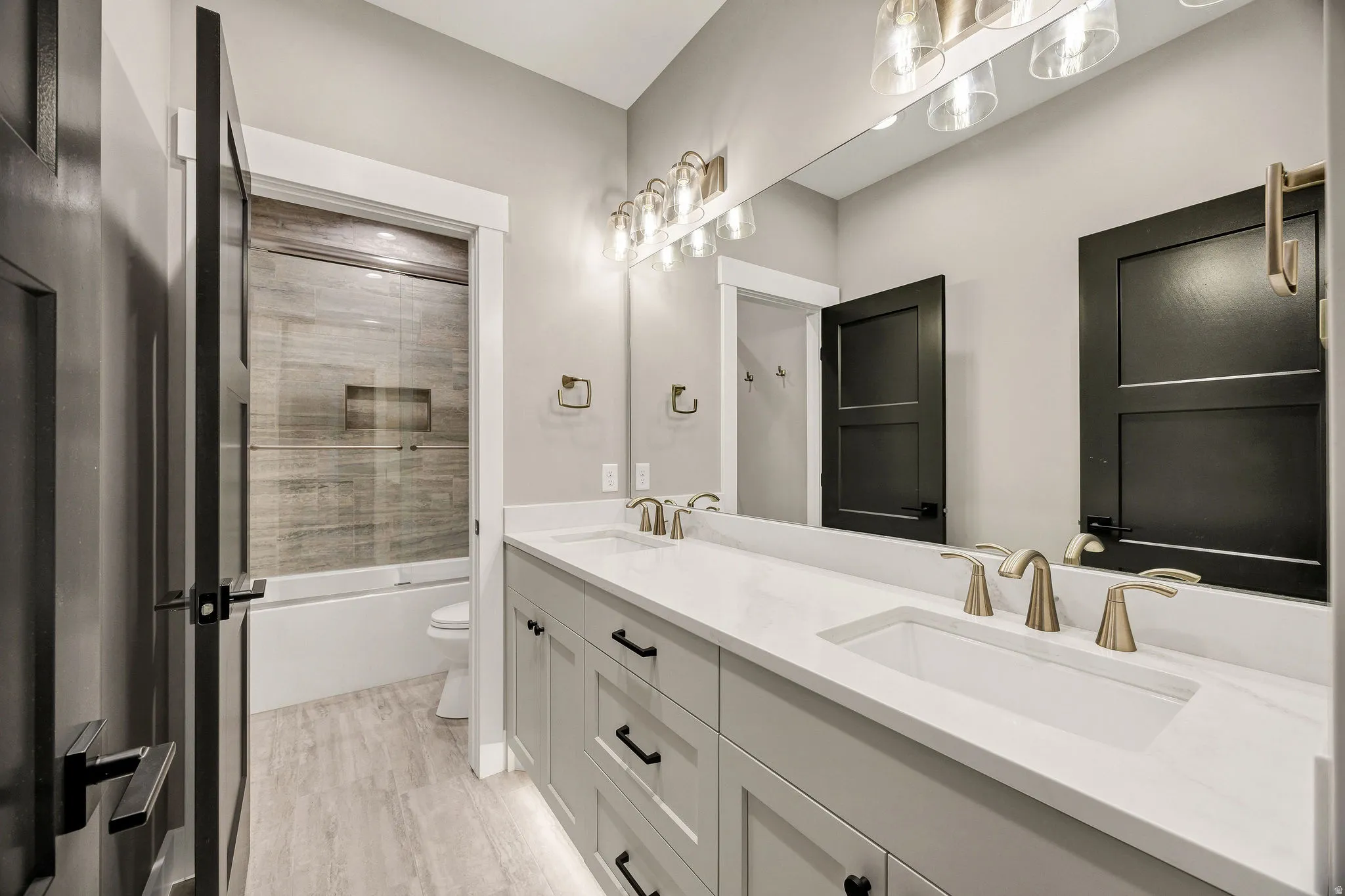 Dual vanity with undermount sinks, light wood-finish flooring, built-in bathtub with glass sliding doors, wood-look tile shower surround, and a long mirrored wall