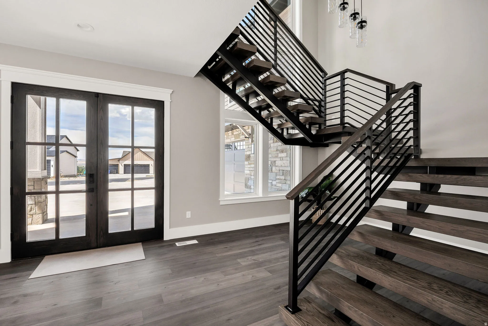 Wood-finish flooring, dark-frame French doors, contemporary staircase with horizontal metal balusters, and a modern chandelier