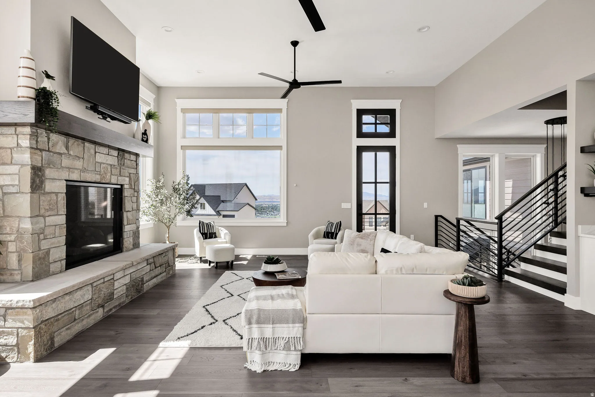 Open-concept living space anchored by a stone fireplace with a black insert, dark wood-finish flooring throughout, and abundant natural light from oversized windows