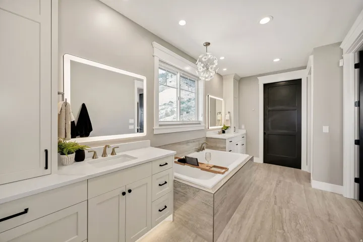 Dual vanity with undermount sinks and light-colored countertops, illuminated mirrors, built-in soaking tub, wood-finish flooring, and a modern chandelier