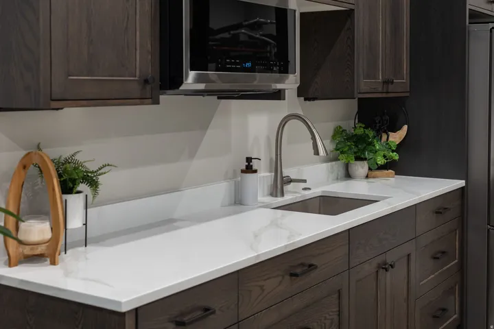 Dark wood-finish cabinetry, stainless steel microwave, white countertop, undermount sink, and brushed nickel faucet