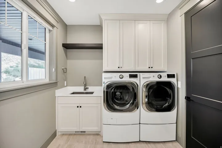 Laundry room featuring white cabinetry, a utility sink with a pull-down faucet, a dark wood-finish floating shelf, and light wood-finish flooring
