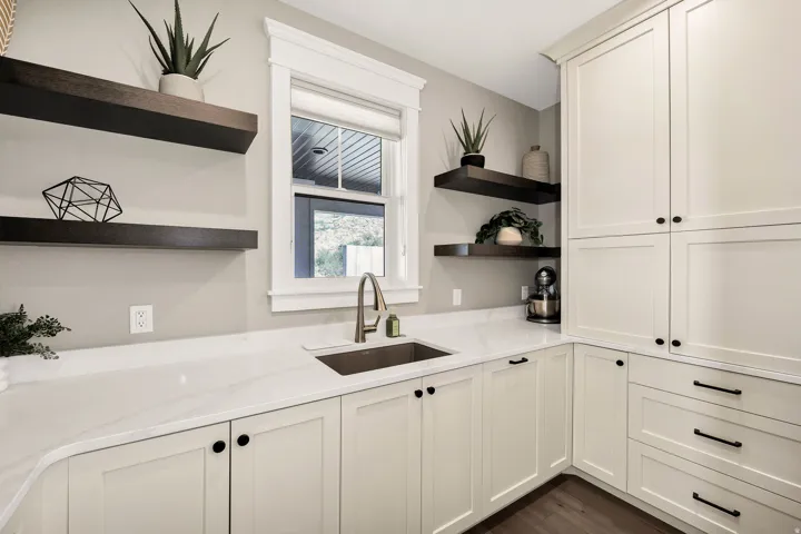 White shaker-style cabinetry with matte black hardware, white countertops, stainless steel sink with a gooseneck faucet, dark wood-finish open shelving, and dark wood-finish flooring