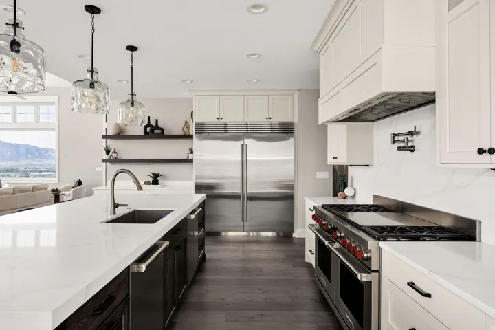 White shaker cabinetry, stainless steel appliances, dark wood-finish flooring, island with sink, and pendulum lighting