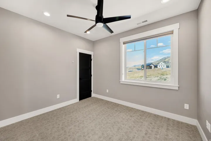 Bright bedroom showcasing neutral wall finishes, carpeted flooring, a contemporary black ceiling fan with integrated lighting, and expansive windows framing natural light and expansive outdoor vistas