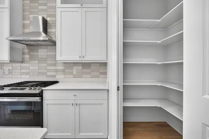 White shaker cabinetry, stainless steel range with overhead hood, light-toned tiled backsplash, white countertops, and corner pantry with ample shelving