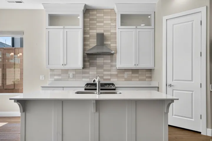 Kitchen featuring a gas range, stainless steel range hood, white shaker cabinets with glass accents, light-toned tiled backsplash, and a central island with an undermount sink and faucet