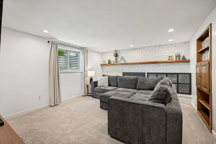 Recessed ceiling lighting, light-colored carpeting, white painted brick accent wall, wood-finish floating shelf, and built-in wood-finish shelving