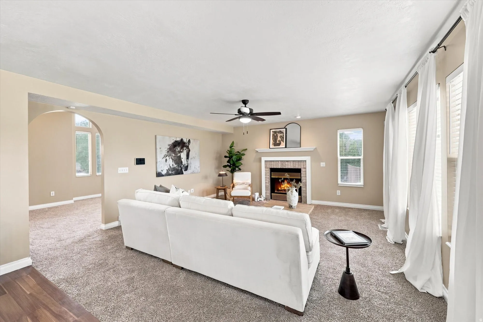 Spacious living area featuring a central fireplace with brick surround and mantle, multiple windows with white blinds, and a ceiling fan