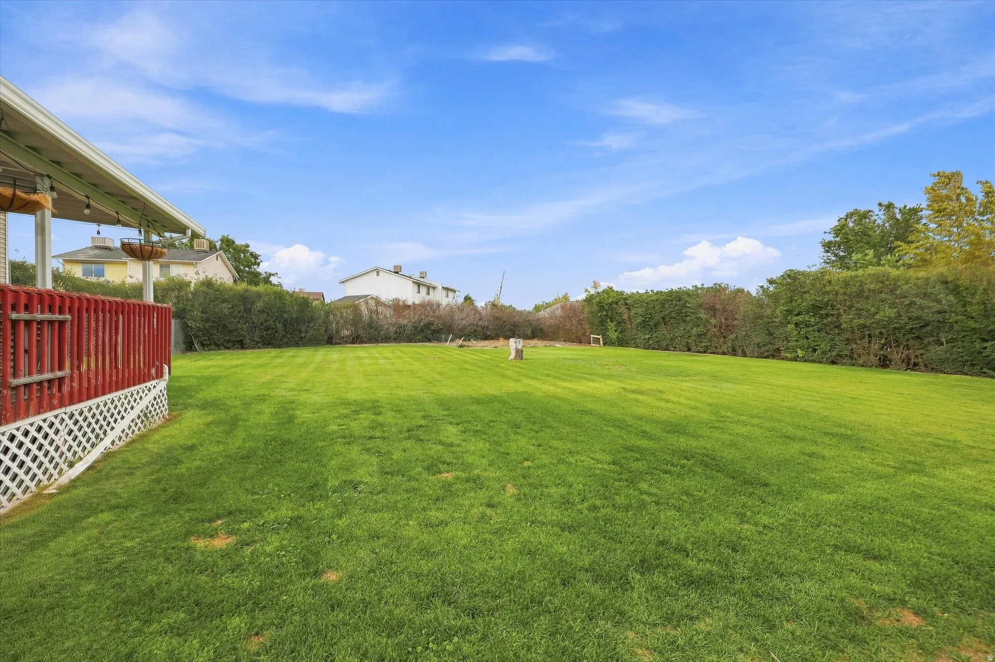 Expansive green lawn, mature perimeter landscaping, covered patio with string lighting, and red-painted wooden deck with lattice detailing