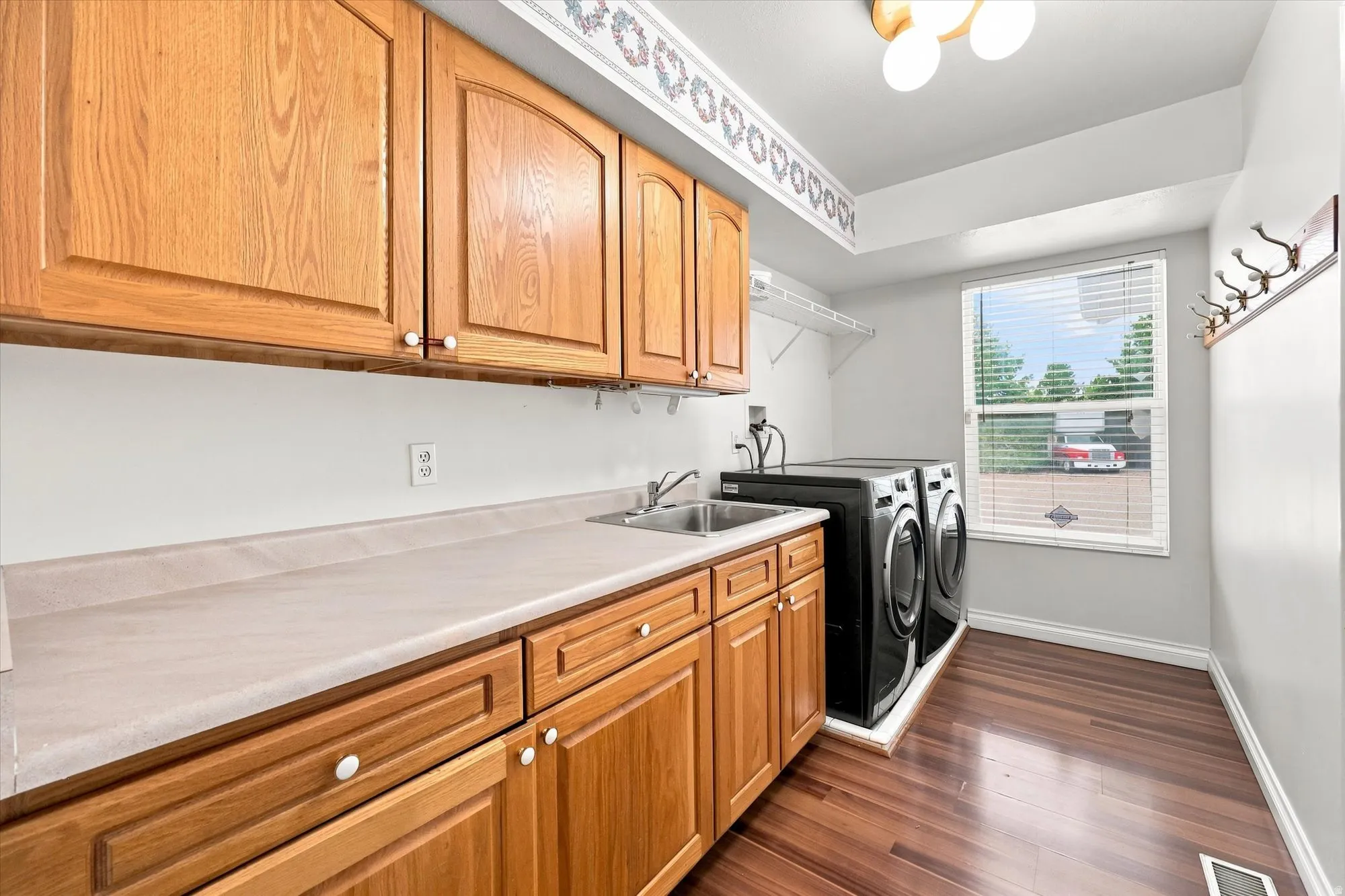Wood-finish flooring, light wood cabinetry, dual stainless steel sink, light-toned countertop, and a window with blinds