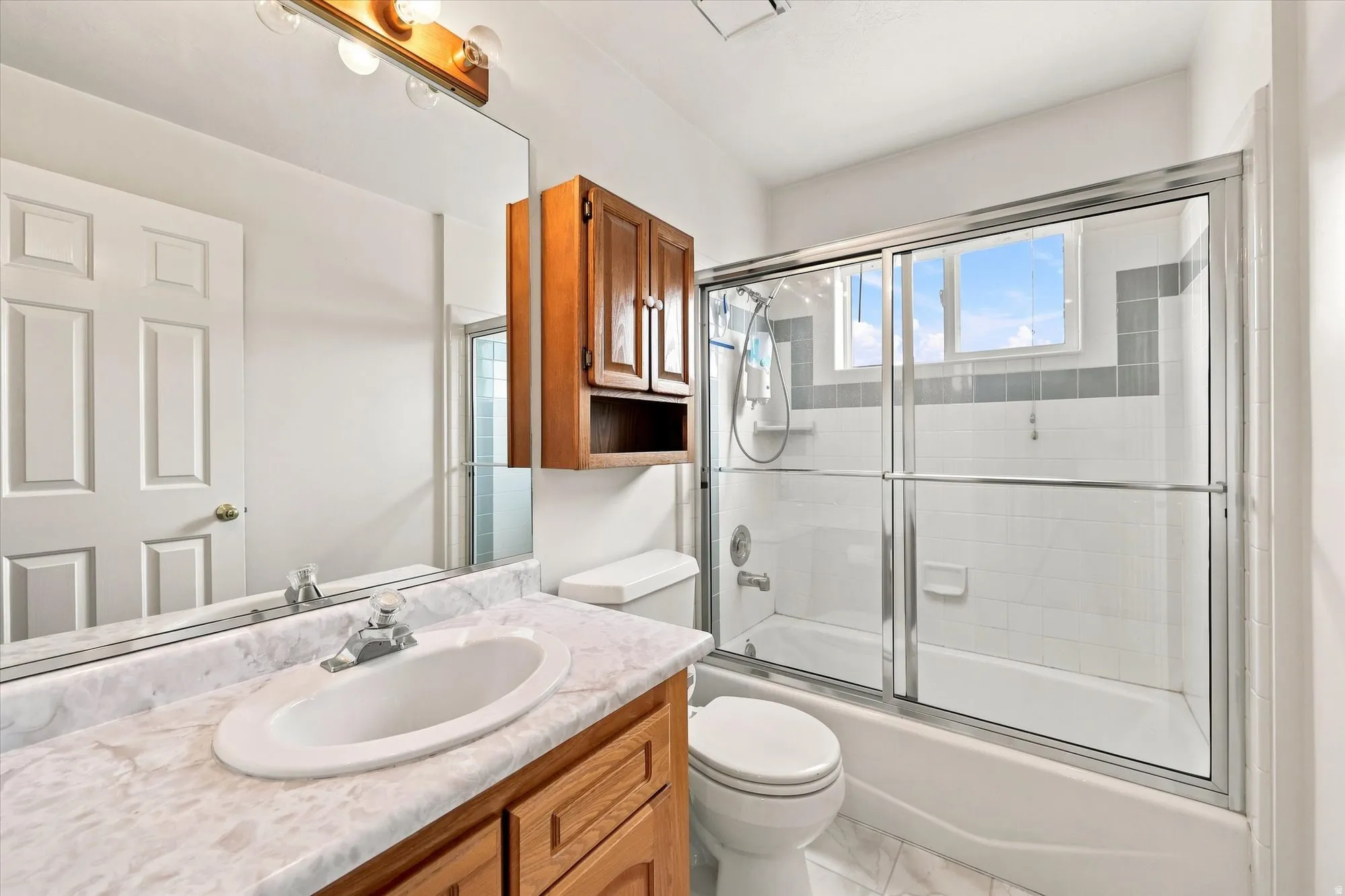 Bathroom featuring a wood vanity with a white countertop and integrated sink, an overhead wood cabinet, a full-width mirror, and a shower-tub combination with sliding glass doors