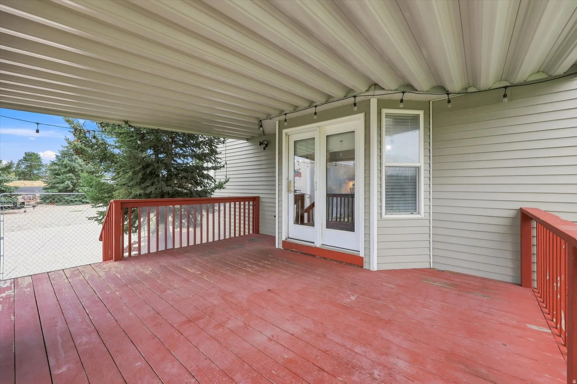 Covered wooden deck with red stained flooring, white double doors, and light gray siding