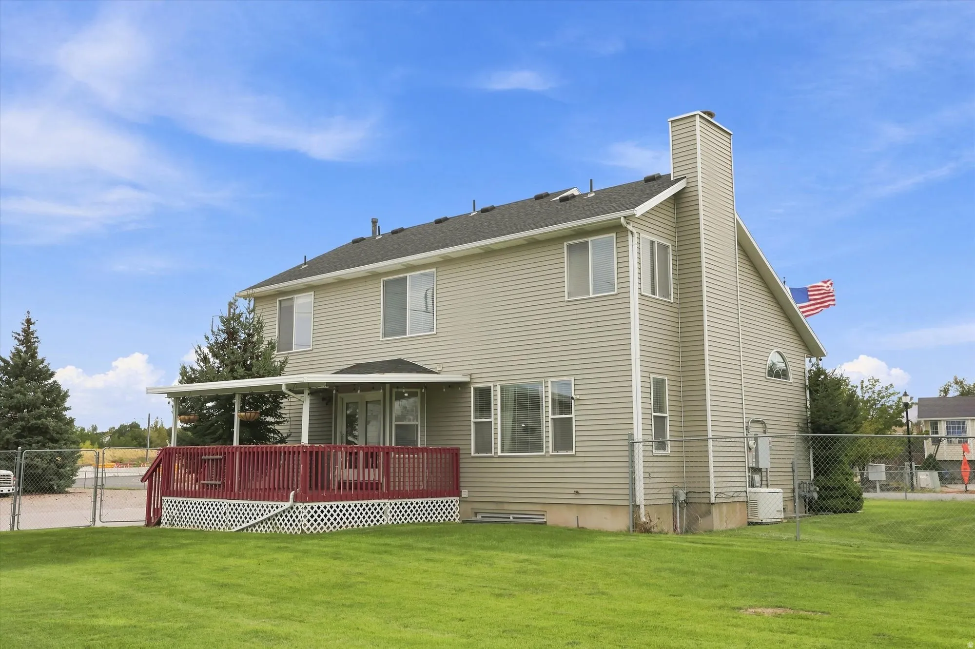 Expansive green space featuring a covered deck with red railings and white lattice, light-colored siding, and a prominent chimney