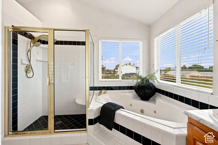 Corner jetted tub with tiled surround, walk-in shower with gold-tone frame, dual windows with blinds, white tile flooring, and a wood-finish vanity with a stone countertop