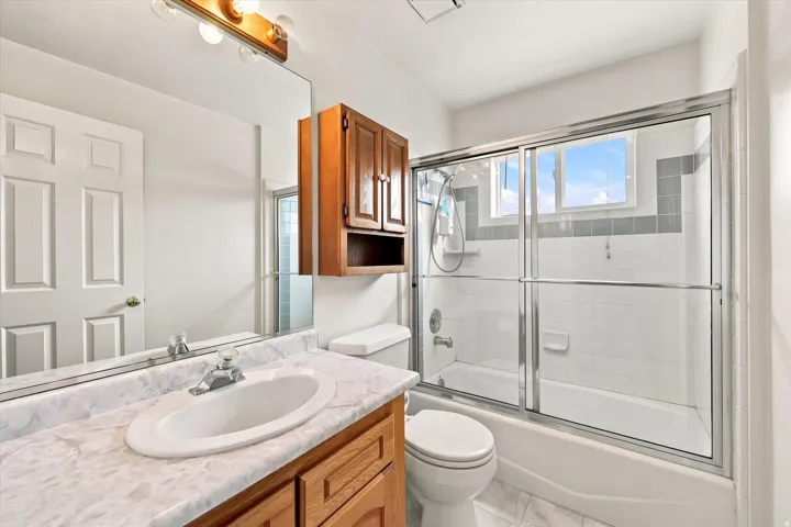 Bathroom featuring a wood vanity with a white countertop and integrated sink, an overhead wood cabinet, a full-width mirror, and a shower-tub combination with sliding glass doors