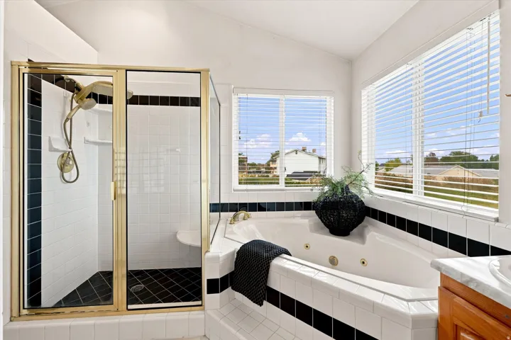 Corner jetted tub with tiled surround, walk-in shower with gold-tone frame, dual windows with blinds, white tile flooring, and a wood-finish vanity with a stone countertop