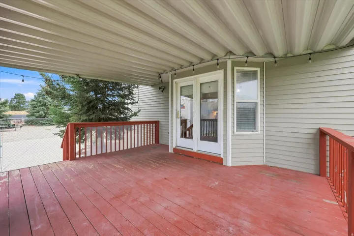 Covered wooden deck with red stained flooring, white double doors, and light gray siding