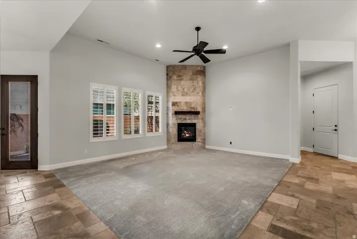 Vaulted ceiling with an attached ceiling fan, floor-to-ceiling stone fireplace, three windows with plantation shutters, light gray carpet, and stone-finish tile flooring