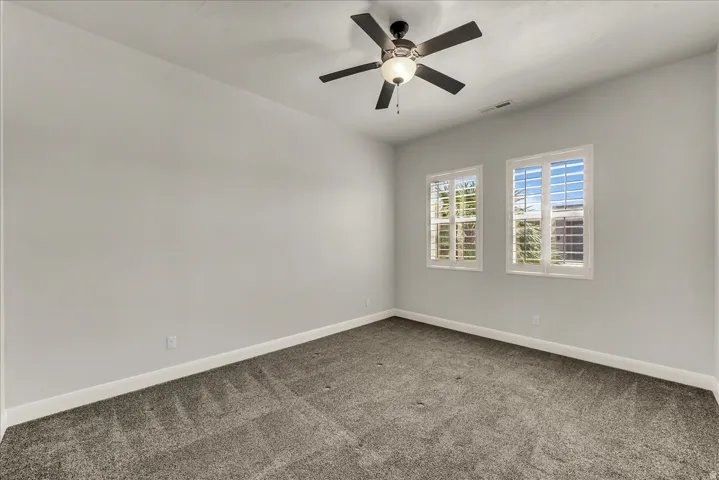 Soft gray wall color, two windows with plantation shutters, ceiling fan with integrated lighting, and neutral carpet flooring
