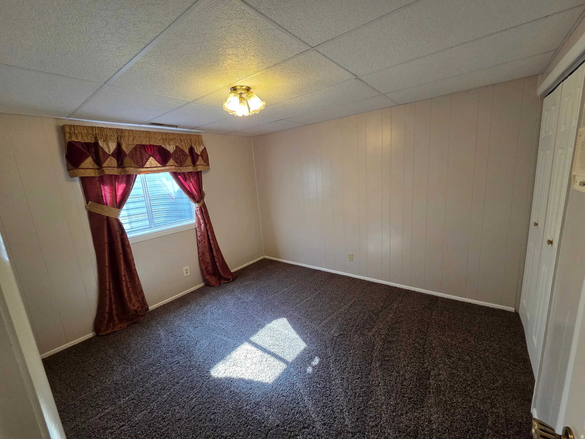 Textured ceiling panels, wall paneling, window with blinds, bi-fold closet doors, and a ceiling-mounted light fixture
