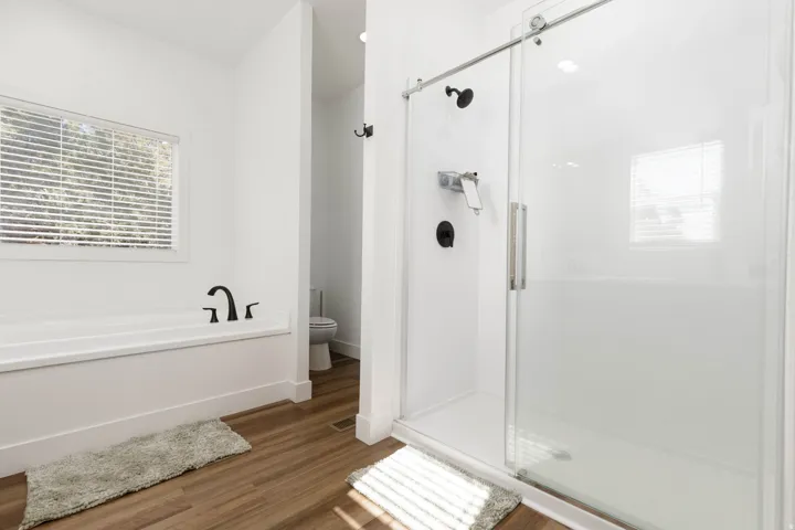 Glass-enclosed shower with matte black fixtures, separate soaking tub with matte black faucet, wood-finish flooring, and bright white walls