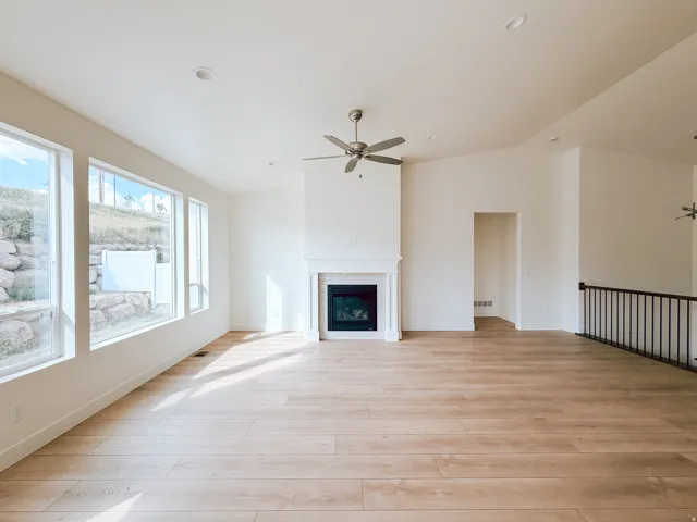 Spacious living area featuring wood-finish flooring, a fireplace with a mantel, multiple large windows, and a ceiling fan