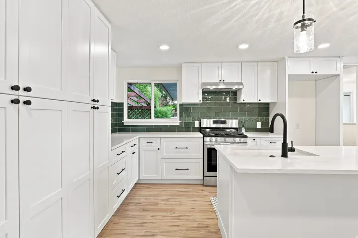 White shaker-style cabinetry with matte black hardware, stainless steel range and hood, green subway tile backsplash, and light wood-finish flooring