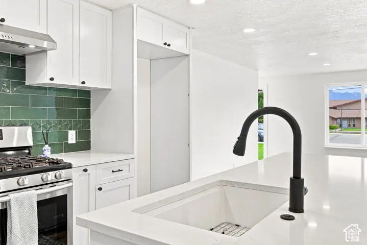 White cabinetry, bright green tile backsplash, stainless steel range with hood, white countertop, and a built-in white sink with a matte black faucet