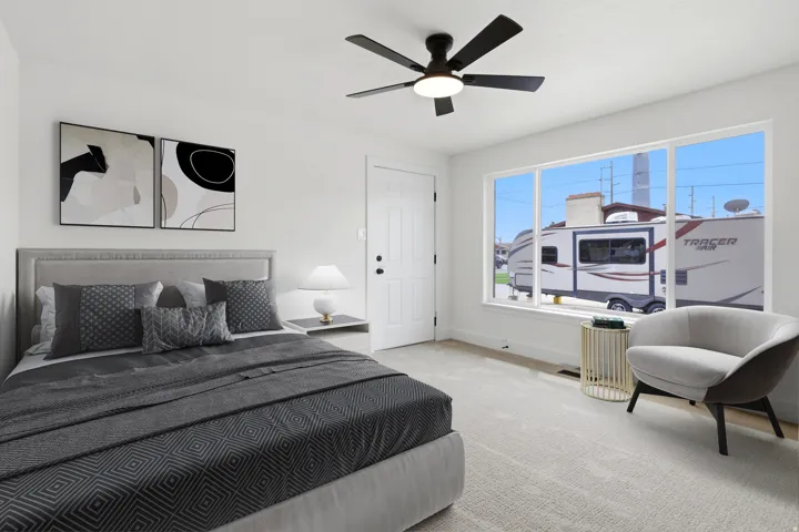 Master bedroom ceiling fan lighting, broad window, paneled door, and neutral wall color with plush carpet.