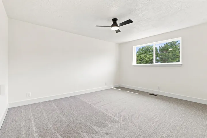 Spacious interior with neutral wall tones, light gray patterned carpeting, a large window, and a ceiling fan with integrated lighting
